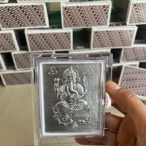 May include: A rectangular silver-coloured plaque featuring an embossed image of the deity Ganesha seated in a cross-legged position. The plaque includes the Om symbol on both sides and is encased in a clear, transparent acrylic protective frame.