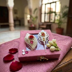 Puede incluir: Una caja de terciopelo rosa que contiene un adorno plateado en forma de hoja con una rosa roja y una gema rosa, y un broche plateado en forma de pluma con una gema roja. La caja está decorada con flores de jazmín blancas y pétalos de rosa.