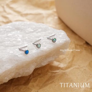 May include: Three titanium nose studs with opal stones in blue and green hues. The studs are displayed on a white surface against a brown backdrop. Text on the image reads "2.5mm", "20g(0.8mm)/7mm", and "TITANIUM".