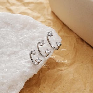 Könnte beinhalten: Drei silberfarbene Nasenringe mit klaren, funkelnden Edelsteinen. Jeder Ring hat ein gebogenes Design mit einem Edelstein an jedem Ende. Größen: 7mm, 8mm und 9mm. Stärke: 18g (1,0mm).