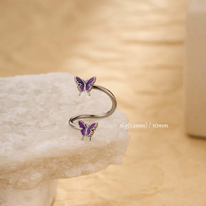 May include: A silver-colored, curved body jewelry piece with two purple butterfly accents. The butterflies have detailed wing patterns. The text "16g (1.2mm) / 10mm" is visible on the right side of the image.