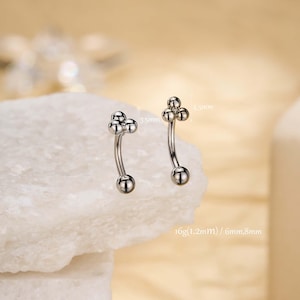 Könnte beinhalten: Zwei silberfarbene, gebogene Augenbrauenpiercings. Jeder Ring hat an einem Ende eine Ansammlung von drei kleinen Kugeln und am anderen Ende eine einzelne Kugel. Maße: 3,5 mm und 4,5 mm, und 16g (1,2 mm) / 6 mm, 8 mm.