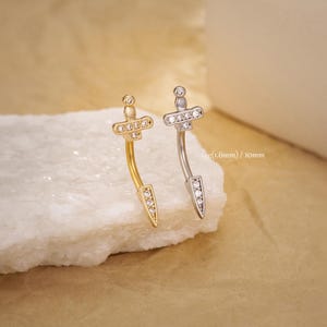 May include: Two curved belly button rings, one gold-toned and one silver-toned, each featuring a dagger design. The daggers have a cross-shaped top and a pointed bottom, both adorned with clear gemstones. The text "16g(1.6mm) / 10mm" is visible.
