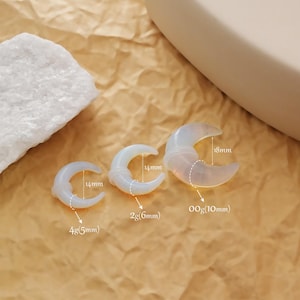 May include: Three crescent moon-shaped ear gauges in varying sizes. The gauges are white and translucent, with measurements of 14mm, 18mm, 6mm, 10mm, and 5mm. The gauges are displayed on a textured, tan surface.