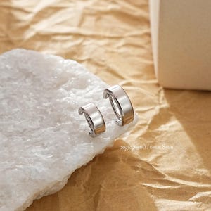 May include: A pair of silver hoop earrings, one slightly larger than the other, are presented on a white, textured surface. The earrings are set against a brown, textured backdrop. The image text reads: 20g(0.8mm) / 6mm,8mm.