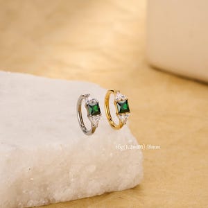 Peut inclure: Deux boucles d'oreilles créoles, une argentée et une dorée, chacune ornée d'une pierre verte émeraude carrée. Les boucles d'oreilles sont ornées de plus petites pierres précieuses claires. Le texte "16g(1.2mm) / 8mm" est visible.