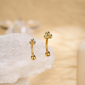 Könnte beinhalten: Zwei goldfarbene Augenbrauen-Piercing-Schmuckstücke. Jedes Stück hat einen gebogenen Stab mit einer kleinen Kugel an jedem Ende und einer Ansammlung von drei kleinen Kugeln oben. Der Schmuck wird auf einer weißen Oberfläche präsentiert.