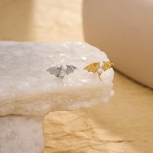 May include: Two stud earrings shaped like small winged creatures with outstretched wings. One earring is silver-toned and the other is gold-toned. Each features a central marquise-cut clear gemstone set in the body, with detailed wing textures.