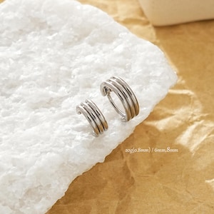 Peut inclure: Boucles d'oreilles créoles argentées avec un design à plusieurs couches, présentées sur une surface blanche texturée sur fond beige. Diamètre de 6 mm et 8 mm. Texte : 20g(0.8mm) / 6mm,8mm.