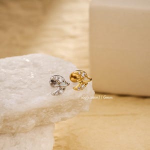 May include: Two earrings, one silver and one gold, each featuring a crescent moon design encrusted with small, clear gemstones. The earrings are displayed on a white, textured surface. Text on the image reads: 16g (1.2mm) / 6mm.