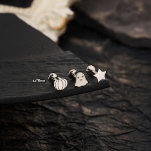 May include: Three silver-toned metal stud earrings featuring a pumpkin, a ghost, and a star shape. Each earring has a flat back post and is labelled with the measurement 1.2*8mm. The jewellery pieces are arranged in a row on a dark textured surface.