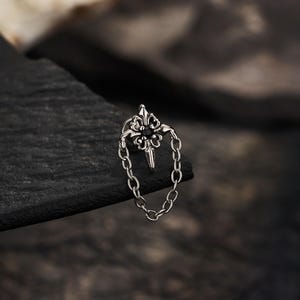 May include: A silver-toned metal stud earring featuring an ornate, symmetrical floral-inspired centrepiece with a small black stone at its centre. A delicate metal chain is attached to the top and bottom of the centrepiece, forming a loop.