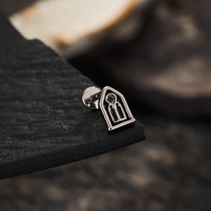 May include: A silver-toned metal stud earring featuring a flat, pointed arch design that resembles a gothic window frame. The piece includes a circular backing post and a cutout detail within the arch shape, finished with a polished metallic surface.