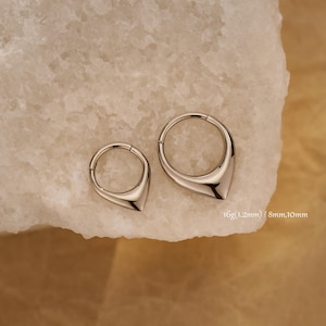 Op de afbeelding: Twee zilverkleurige neusringen in de vorm van een druppel. De ringen worden gepresenteerd op een wit, getextureerd oppervlak. De tekst op de afbeelding luidt: 16g (1,2 mm) / 8 mm, 10 mm.