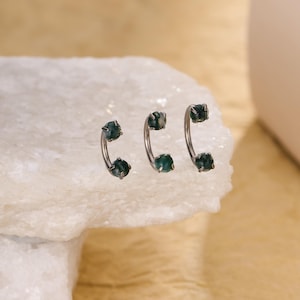 Pode incluir: Três piercings de barra curva com hastes de metal prateado e esferas de pedra verde-escura, semelhantes a musgo, em cada extremidade. As peças de joalharia estão dispostas em fila, exibindo as pedras polidas presas por pequenas garras.