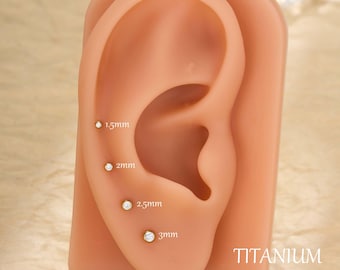 Puede incluir: Exhibición de cuatro pendientes de botón pequeños y redondos, de color dorado, con piedras preciosas transparentes, que varían en tamaño de 1,5 mm a 3 mm. Los pendientes están colocados en un modelo de oreja color carne. La palabra "TITANIUM" está impresa en blanco.