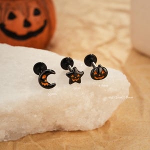 May include: Black Halloween-themed earrings featuring a crescent moon, star, and pumpkin design. Each earring has a black post and a matching orange face. The moon earring is 5.5mm, the star is 5mm, and the pumpkin is 4-8mm.