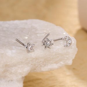 Puede incluir: Tres piercings nasales plateados con diseños celestiales. Uno tiene forma de estrella, otro de sol y el tercero de media luna. Cada uno está adornado con pequeñas piedras preciosas transparentes. El texto "20g/7mm" es visible.