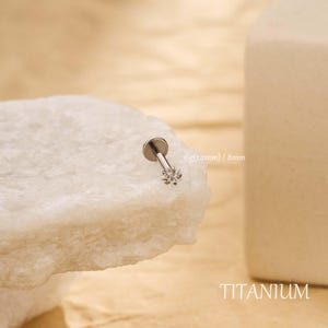Puede incluir: Un pendiente labret de titanio plateado con un diseño en forma de flor. El pendiente mide 8 mm de largo y 1,2 mm de grosor. La palabra "TITANIUM" está impresa en la parte inferior de la imagen.