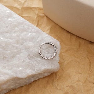 May include: A silver-colored, hinged nose ring with a twisted rope design. The ring is circular and features a continuous pattern of interwoven metal. The jewelry is displayed on a white, textured surface against a neutral background.