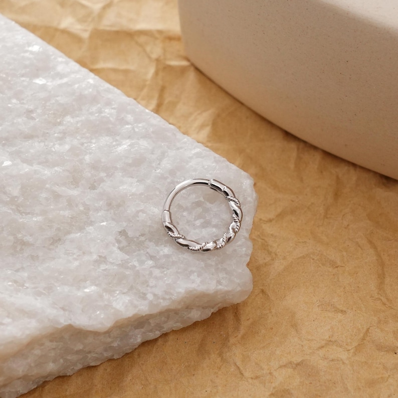 May include: A silver-colored, hinged nose ring with a twisted rope design. The ring is circular and features a continuous pattern of interwoven metal. The jewelry is displayed on a white, textured surface against a neutral background.