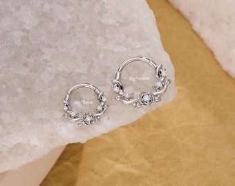 May include: Two silver-colored nose rings with floral and gemstone accents. The rings are circular, with a hinged design. One ring is labeled "16g/8mm" and the other "16g/10mm". The jewelry is displayed on a white and gold surface.