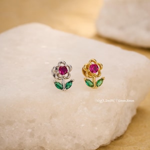 May include: A pair of flower-shaped stud earrings. One earring is silver-toned, the other gold-toned. Each has a pink gemstone center, green leaf accents, and measures 6mm by 8mm.