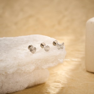 Puede incluir: Tres pendientes de botón plateados con diferentes diseños: un sol, un círculo y una mariposa. Los pendientes se muestran sobre una superficie blanca texturizada. El texto "16g (1.2mm) / 8mm" es visible.