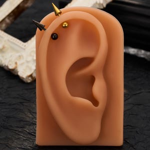 Op de afbeelding: Een siliconen oormodel met vier piercings langs het bovenste kraakbeen. De sieraden bestaan uit twee metalen stekers in de vorm van een punt in goud en zwart, gecombineerd met twee gladde bolvormige stekers in bijpassende kleuren.