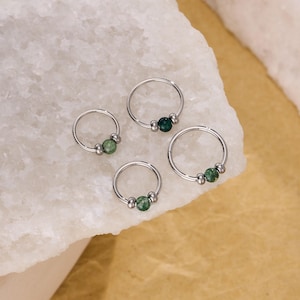 May include: Four silver-colored hoop earrings with green gemstone beads. The earrings are arranged on a white and gold surface. The gemstones are a deep green color, and the metal has a shiny finish.
