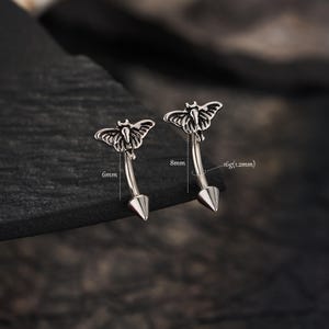 Può includere: Due piercing a barra curva color argento con una parte superiore a forma di falena e una base a cono appuntito. Il pezzo a sinistra è etichettato 6mm, mentre quello a destra indica 8mm e 16g (1,2mm) per la lunghezza e lo spessore.