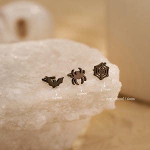 Op de afbeelding: Drie zwarte Halloween-thema oorknopjes: een vleermuis (3,4 mm), een spin (4,9 mm) en een spinnenweb (4,7 mm). De oorbellen worden gepresenteerd op een wit, getextureerd oppervlak. De oorbellen zijn klein en gedetailleerd.