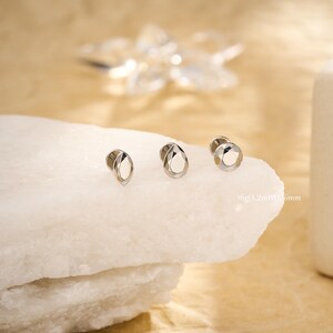 May include: Three silver-colored ear plugs with faceted, teardrop-shaped and round designs. The plugs are displayed on a white, textured surface. The text "10g (1.2mm) 6mm" is visible in the image.