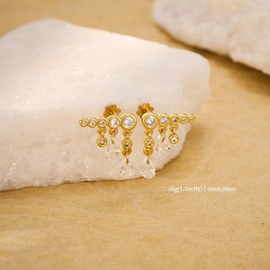 May include: Gold-toned earrings with a series of small, round, clear gemstones and dangling teardrop-shaped gems. The earrings are displayed on a white, textured surface. Text on the image reads: 16g(1.2mm) / 6mm,8mm.