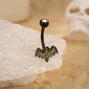 May include: Black belly button ring with a bat-shaped charm. The bat is decorated with green and pink glitter. The top ball has a clear gem. The ring is 10mm long and the bat charm is 8.3mm wide.
