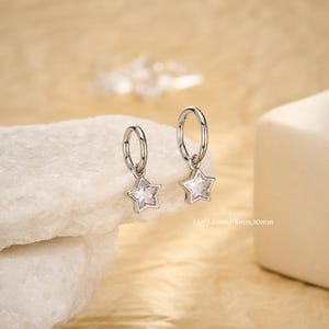 May include: A pair of silver-colored hoop earrings with star-shaped pendants. Each star has a clear, faceted center. The earrings are displayed on a white, textured surface against a soft, neutral background. Text on the image reads: 14g(1.6mm)/8mm,10mm.