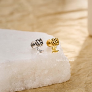 Puede incluir: Dos pendientes con forma de rosa, uno plateado y otro dorado, se muestran sobre una superficie blanca. Los pendientes tienen pétalos y hojas detallados. El texto de la imagen dice "16g (1,2 mm) / 8 mm".