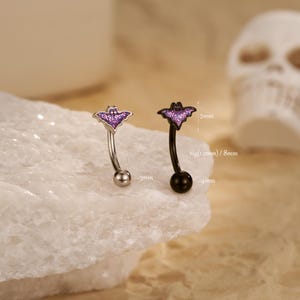 May include: Two curved eyebrow rings with bat-shaped charms. One ring is silver with a purple bat, the other is black with a purple bat. The bat charms are approximately 5mm wide. The silver ring is 3mm wide, and the black ring is 4mm wide.