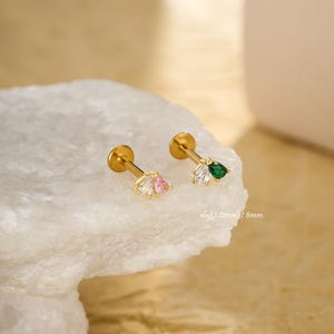 Könnte beinhalten: Zwei goldfarbene Ohrstecker mit dekorativen Enden. Ein Stecker hat einen Cluster aus klaren und rosa birnenförmigen Edelsteinen, der andere einen Cluster aus klaren und grünen Edelsteinen. Die Stecker sind auf einer weißen, strukturierten Oberfläche.
