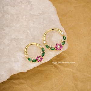 Peut inclure: Deux boucles d'oreilles dorées en forme de cercle. Chaque boucle d'oreille présente un motif floral avec des pierres précieuses vertes et roses, et de petits gemmes blancs. Les boucles d'oreilles sont exposées sur une surface blanche texturée. Texte sur l'image : 16g(1.2mm) / 8mm,10mm.