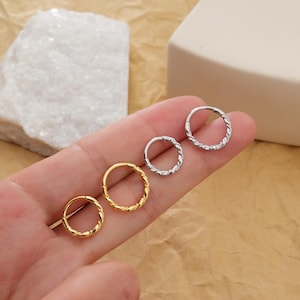 May include: Four small hoop earrings with a twisted rope design. Two are gold-colored, and two are silver-colored. The earrings are displayed on a light brown surface.