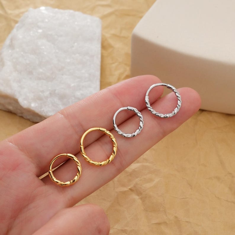 May include: Four small hoop earrings with a twisted rope design. Two are gold-colored, and two are silver-colored. The earrings are displayed on a light brown surface.