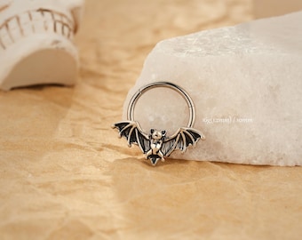 Puede incluir: Un anillo articulado plateado con un dije de murciélago. El murciélago tiene alas detalladas y una pequeña pajarita. El texto "16g (1,2 mm) / 10 mm" es visible. El fondo incluye una piedra blanca y una calavera.