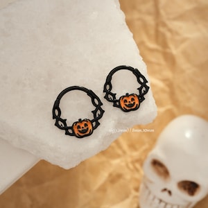 May include: Two black Halloween-themed hoop earrings. Each earring features a smiling orange pumpkin charm. The earrings are displayed on a white surface. The text on the image reads "16g(1.2mm) / 8mm, 10mm".