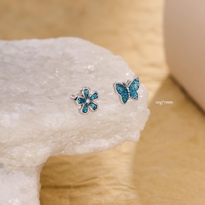Pode incluir: Dois brincos prateados, um em forma de flor e outro de borboleta. Ambos os brincos têm um preenchimento de esmalte azul turquesa. O texto "20g/7mm" está impresso no lado direito da imagem.