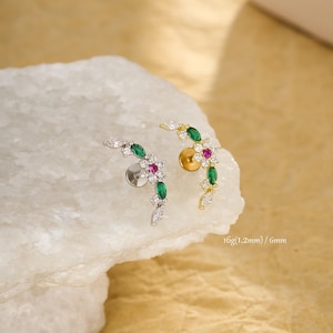 May include: A pair of earrings with a floral design. One earring is silver-toned, the other gold-toned. Each features a cluster of small, white, marquise-cut stones, green emerald-cut stones, and a central pink stone. The text "16g (1.2mm) / 6mm" is visible.