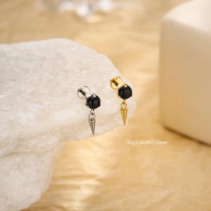 Op de afbeelding: Twee oorstoppers, één zilverkleurig, de andere goudkleurig, met zwarte, bolvormige toppen en hangende, kegelvormige bedels. De tekst "16g (1,2mm) / 6mm" is zichtbaar. De oorstoppers hebben een diameter van 6 mm.