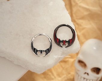 Puede incluir: Dos anillos de septum con forma de murciélago. Uno es plateado con detalles negros, el otro es negro con detalles rojos. Los anillos se muestran sobre una superficie blanca. Texto en la imagen: 16g (1,2 mm) / 10 mm.