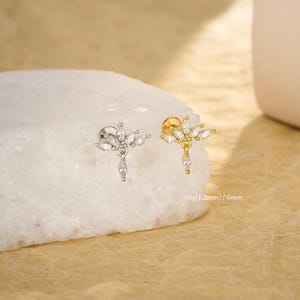 Puede incluir: Dos pendientes de botón, uno plateado y otro dorado, cada uno con un diseño floral con gemas claras talladas en marquesa. Una gema en forma de lágrima cuelga del centro. El texto "16g(1.2mm) / 6mm" es visible.