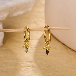 Könnte beinhalten: Ein Paar goldfarbene Creolen mit einem dekorativen Anhänger. Jeder Ohrring hat einen kleinen, grünen Edelstein und ein verziertes, goldfarbenes Design. Die Ohrringe werden auf einer hellen Oberfläche präsentiert.